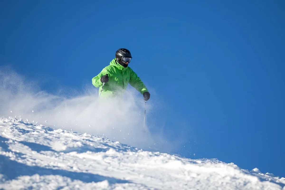 An energetic scene featuring a skier in a bright green jacket descending a sunlit, snow-covered slope beneath a clear blue sky. The image radiates excitement and showcases the region's winter sports offerings.