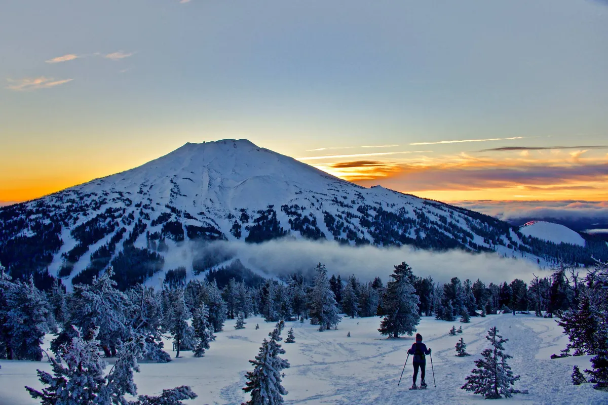 A breathtaking view of a snow-covered mountain with a skier in motion descending the slope, set against a glowing sunset sky and framed by tall pine trees. The scene captures the thrill and beauty of winter sports in a natural alpine environment.