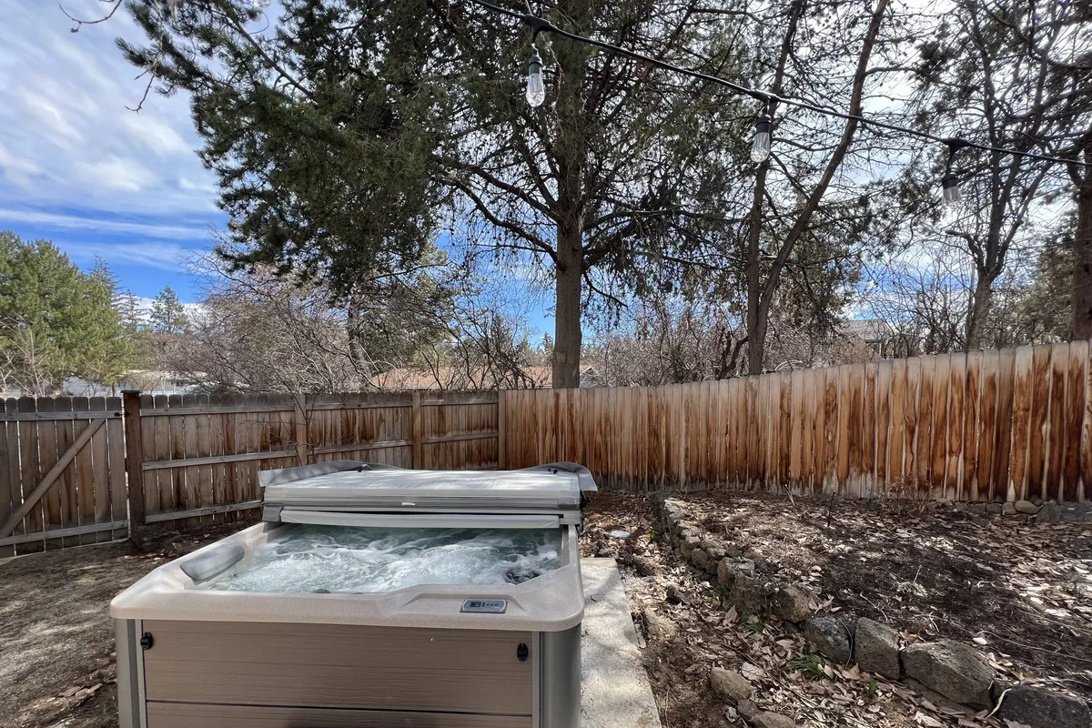 A serene backyard scene featuring a spacious hot tub, bordered by trees and a fence. The setting is ideal for both daytime relaxation and evening gatherings, evoking a sense of comfort and luxury. The image hints at the possibility of barbecues or fireside evenings.