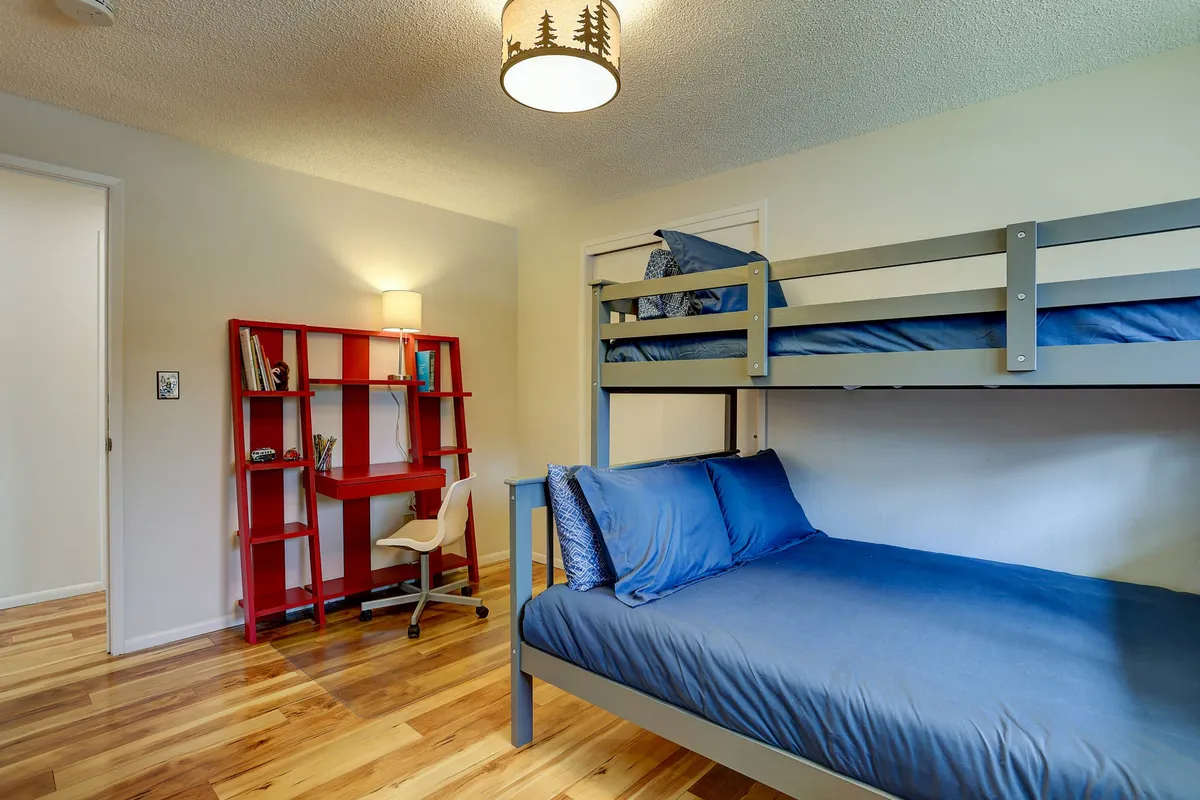 A youthful bedroom with a twin over full bunk bed, a red bookshelf filled with books, and a desk with a chair tucked in, suggesting a flexible space for both rest and study.
