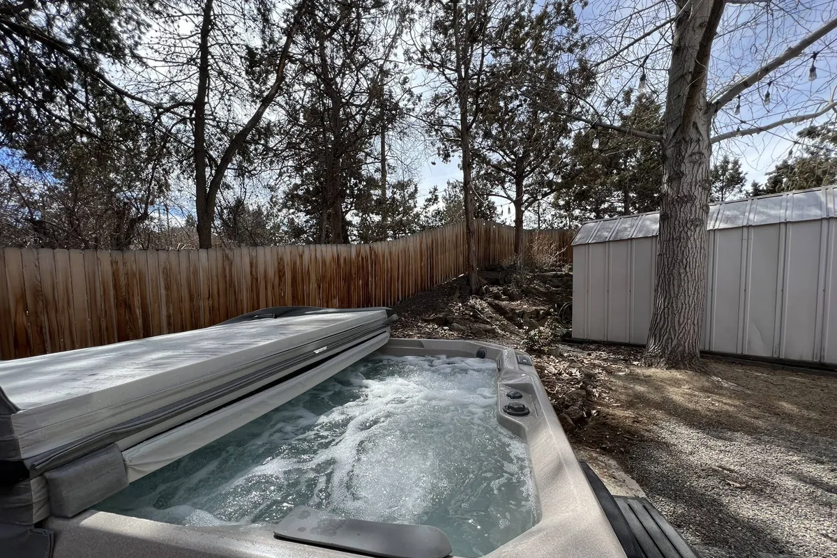 A tranquil backyard setting with a bubbling hot tub surrounded by trees, framed by a secure fenced area. The atmosphere is peaceful and inviting, ideal for relaxation and gatherings. The hot tub is the focal point, offering both privacy and a connection to the outdoors.