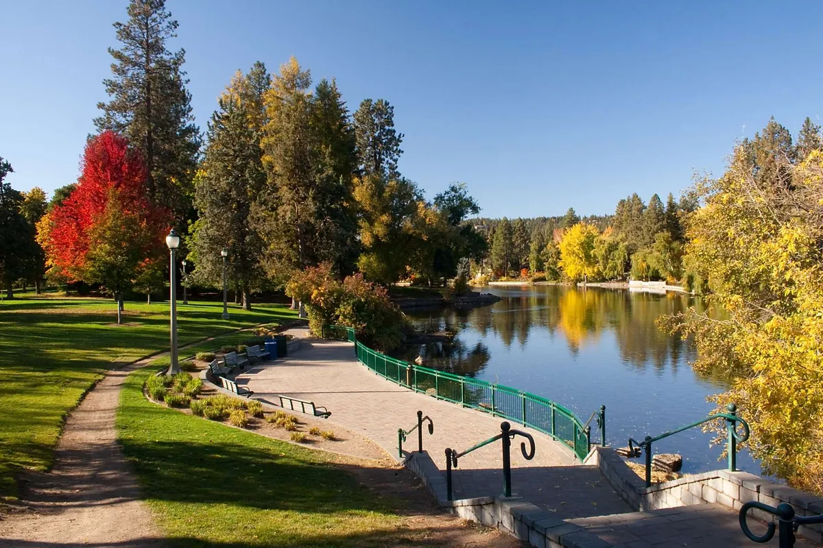 A calm lake bordered by colorful trees, walkways, and park benches, set in a serene, natural environment. The spot is ideal for walking, relaxing, or enjoying seasonal scenery.