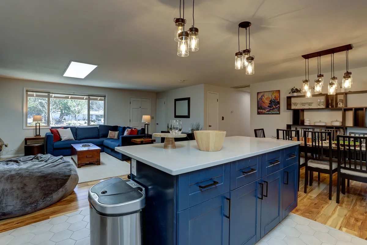 This image captures a vibrant, open-concept interior where the blue sofa and kitchen island visually anchor the space. The frame includes the living area with plush seating and a bowl atop the island, stylish hanging light fixtures, and a view into the dining area. The inviting, cozy ambiance is enhanced by soft natural light and a seamless flow between the living, dining, and kitchen zones.