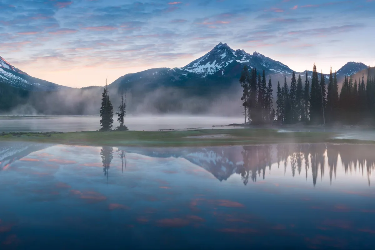 A serene mountain lake bordered by a tree line and towering peaks, with soft early morning light filtering through the clouds. The scene suggests opportunities for hiking, wildlife spotting, and peaceful moments in pristine wilderness.