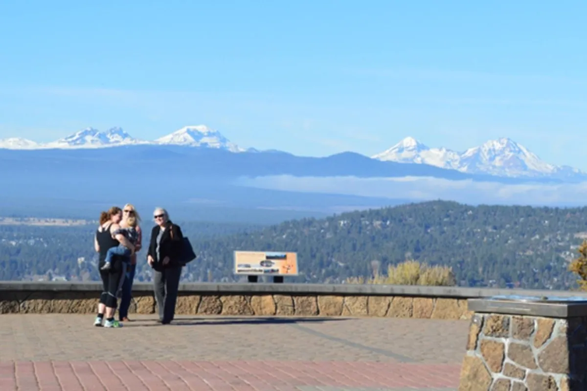 A family group is gathered at a scenic viewing area, with a dramatic mountain range stretching out behind them. The group appears joyful, taking in the panoramic natural beauty.