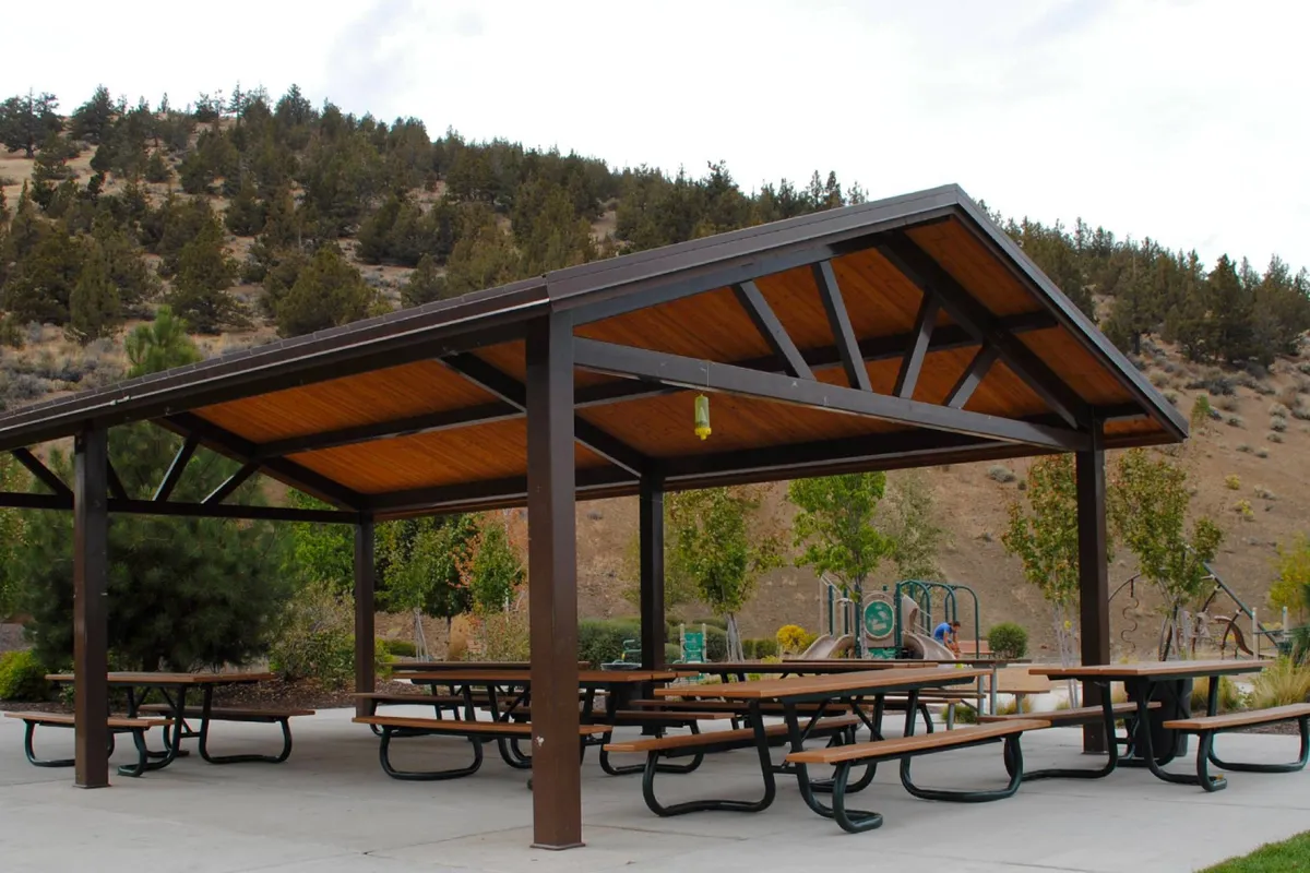 An inviting pavilion set amidst mature trees, benches, and a playground area, with open spaces for picnics and gatherings. Children and families are visible, enjoying the outdoor amenities.