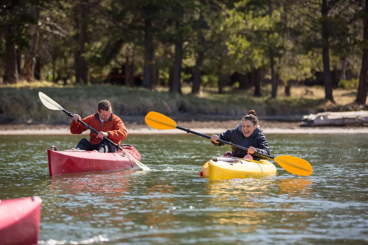 Two kayakers glide across sparkling clear water in vibrant kayaks, surrounded by lush green trees and a tranquil natural landscape. The image radiates a sense of summer fun and outdoor discovery.
