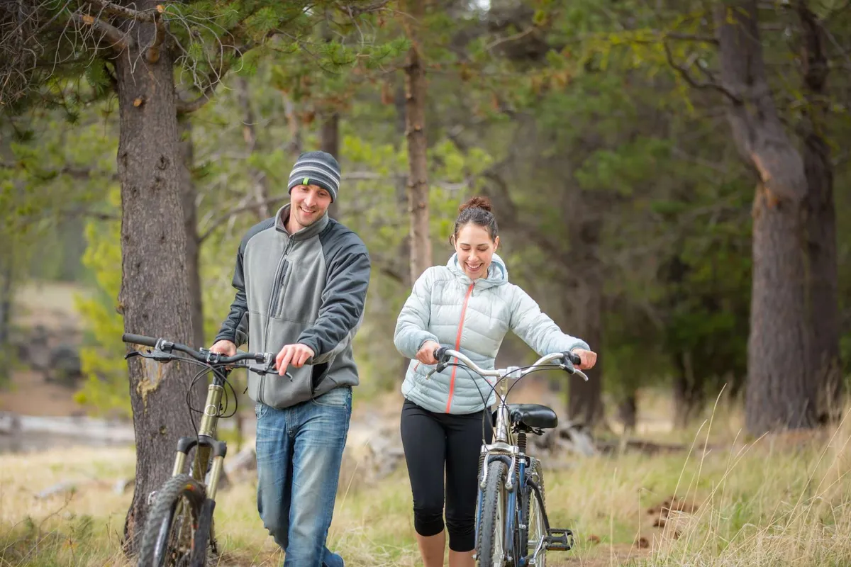 Two cyclists are seen walking with their bikes along a natural pathway bordered by tall trees and lush grassy areas. The background suggests an inviting trail perfect for outdoor exploration, cycling, or hiking.