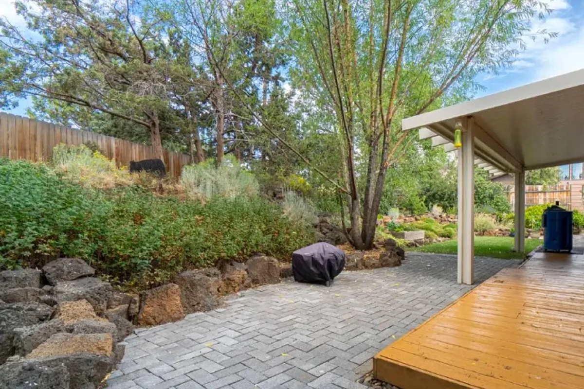 A paved pathway leads through lush vegetation to a wooden deck, suggesting a versatile outdoor space for relaxation, entertaining, or enjoying a morning coffee. The greenery creates a secluded, resort-like feel and hints at the covered patio area.